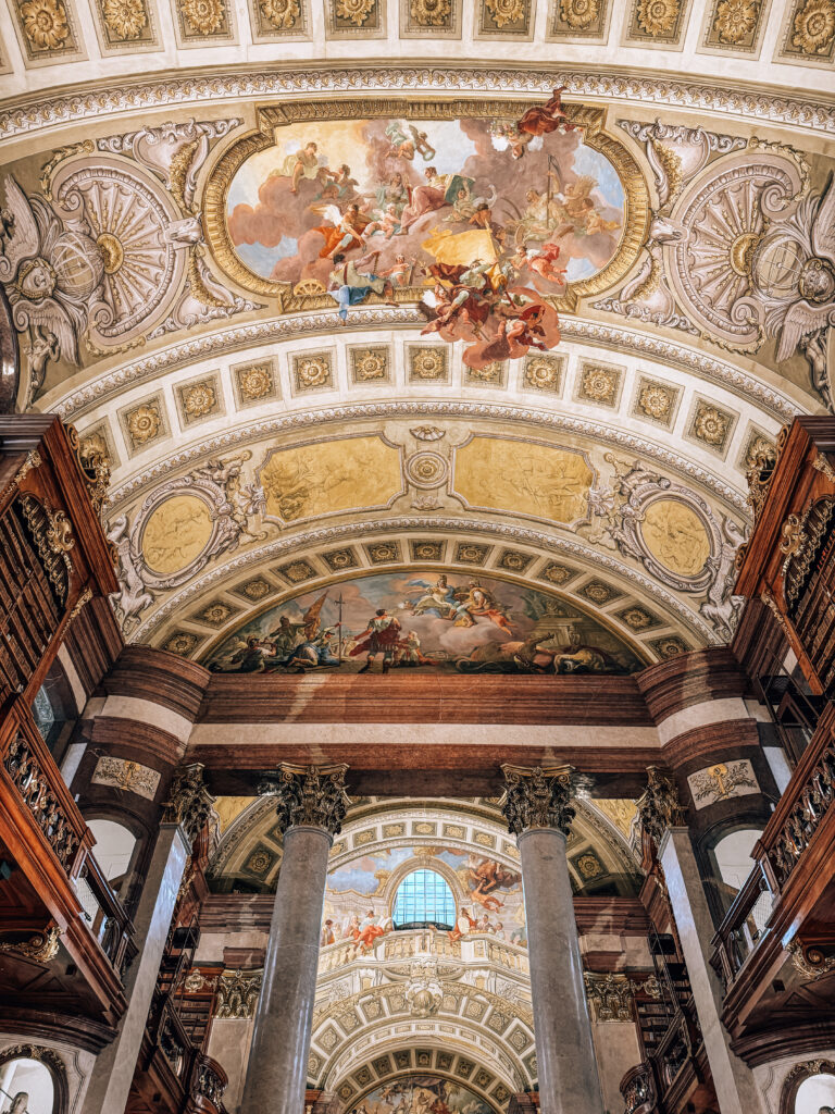 Interior view of the Austrian National Library in Vienna showcasing a grand Baroque hall with marble columns, ornate gold-trimmed balconies, and multiple ceiling frescoes depicting mythological scenes with vibrant figures and clouds.
