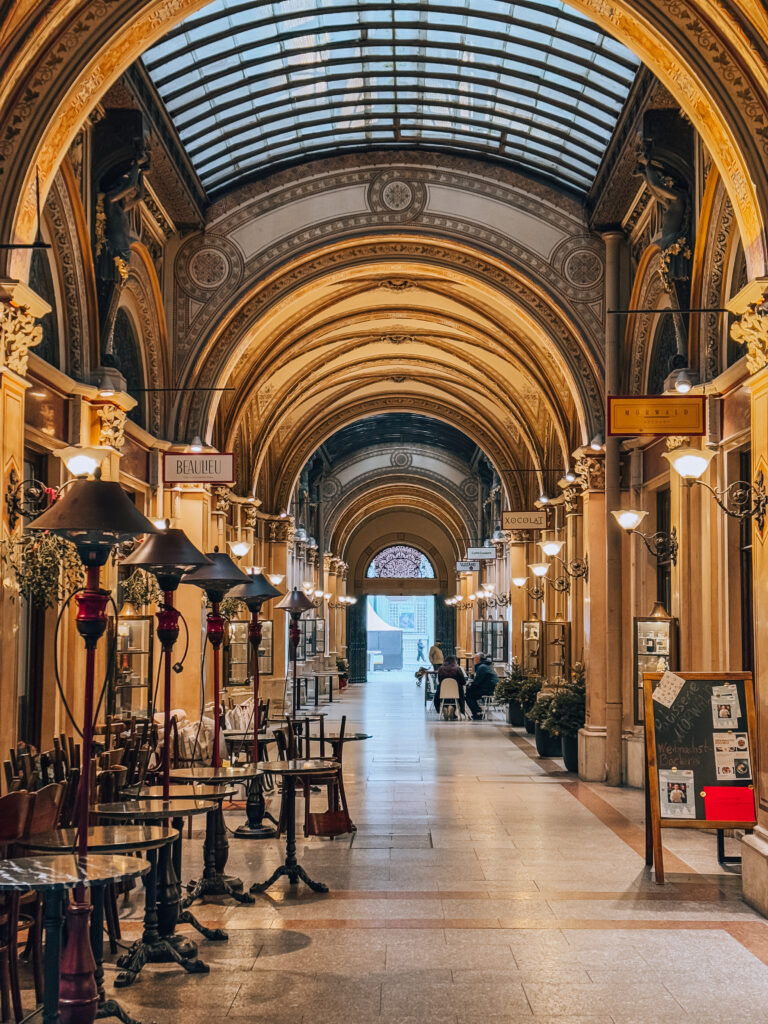 Interior view of the ornate Ferstel Passage in Vienna, featuring a glass-vaulted ceiling, golden arched columns, and elegant café tables lining both sides of the passage.