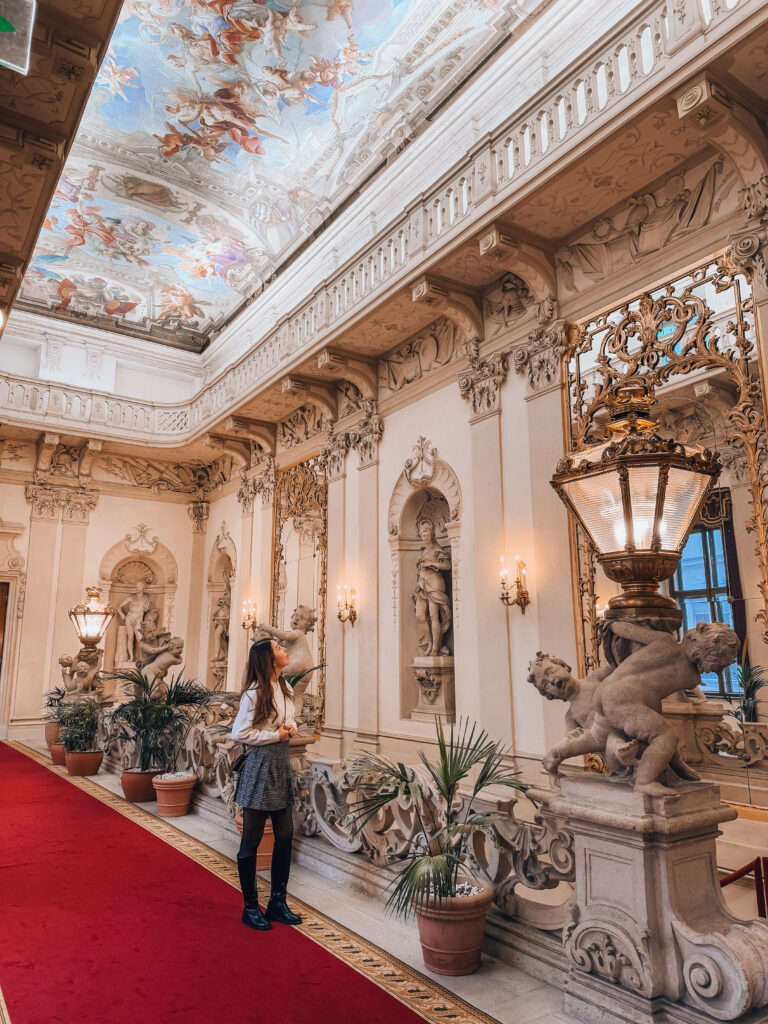 A woman admires the lavish Baroque interior of Palais Daun-Kinsky in Vienna, surrounded by sculpted cherubs, classical statues, ornate lanterns, and a vivid ceiling fresco depicting mythological scenes. The grand hallway is lined with a red carpet and potted palms, highlighting the palace’s opulent elegance.