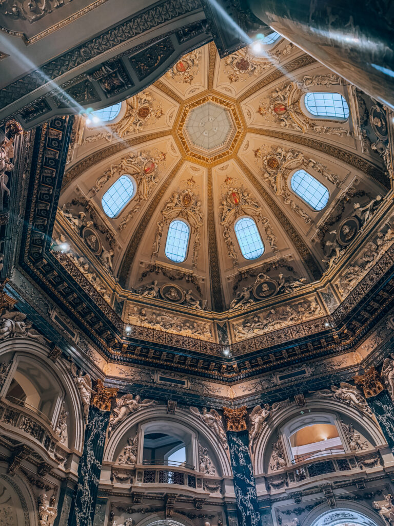 Looking up at the elaborately decorated octagonal dome of the Cupola Hall in the Kunsthistorisches Museum in Vienna, featuring detailed stucco work, classical sculptures, gold accents, and arched windows that let in natural light, creating a majestic atmosphere.
