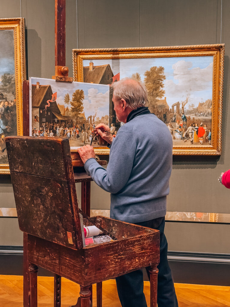 An elderly artist recreates a classical painting inside the Kunsthistorisches Museum in Vienna, seated at a wooden easel with the original framed artwork in the background. The detailed reproduction captures a lively village scene, highlighting the museum’s commitment to art education and preservation.