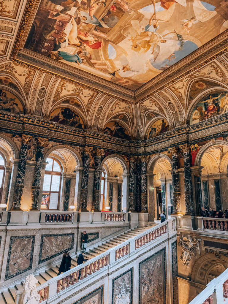 Grand staircase inside the Kunsthistorisches Museum in Vienna, featuring intricately detailed marble walls, arched windows, and a magnificent ceiling fresco with classical figures and vibrant colors. Visitors ascend the stairs beneath the ornate Neo-Renaissance architecture and golden lighting.