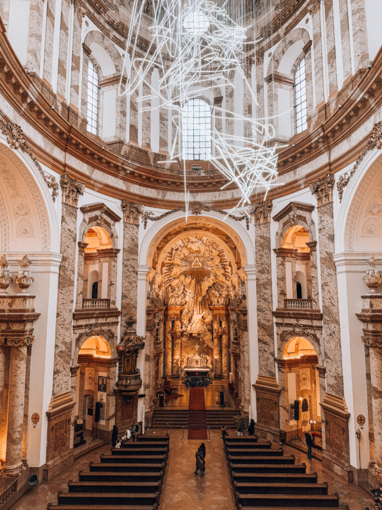 Interior view of Karlskirche in Vienna, showcasing ornate Baroque architecture, grand marble columns, an intricately sculpted golden altar, and a striking modern chandelier installation hanging from the center dome above wooden pews.