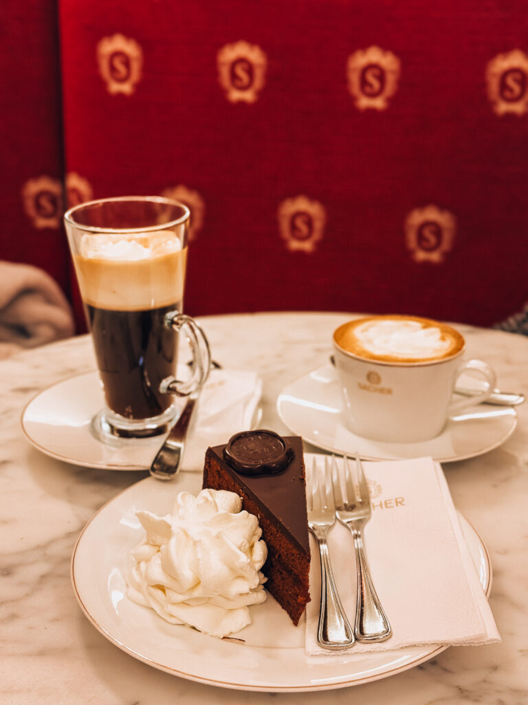 A classic Viennese coffeehouse table setting at Café Sacher featuring a slice of the original Sachertorte with whipped cream, a cappuccino in a branded cup, and a glass of espresso topped with whipped cream, set against a red velvet background embroidered with gold "S" emblems.