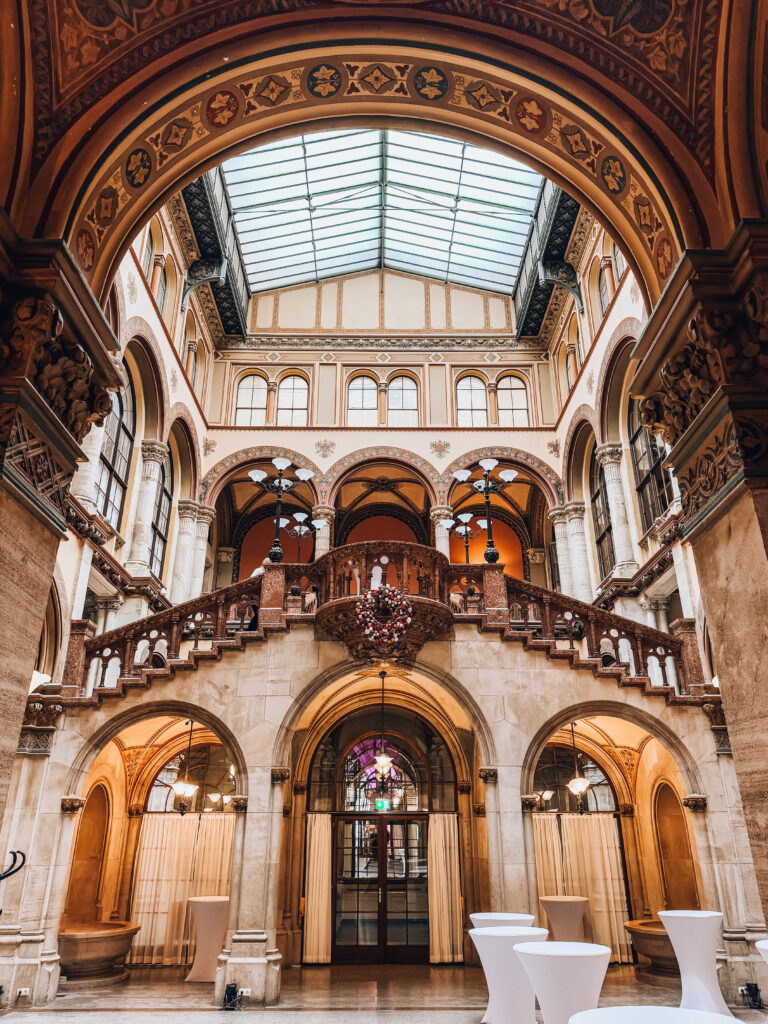 Interior courtyard of Café Central in Vienna, featuring grand arches, a red marble staircase, detailed architectural ornamentation, and a glass-paneled ceiling that fills the space with natural light. Elegant columns and vaulted entryways add to the historic charm of this iconic coffeehouse.