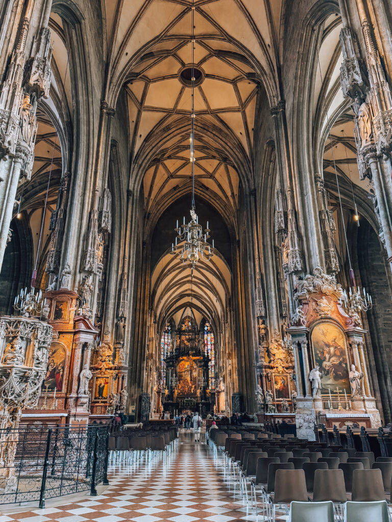 Interior view of St. Stephen’s Cathedral in Vienna, showcasing its soaring Gothic vaulted ceilings, symmetrical stone columns, and richly decorated altars with religious artwork. Rows of chairs line the checkerboard-tiled nave leading to the ornate main altar and stained glass windows.
