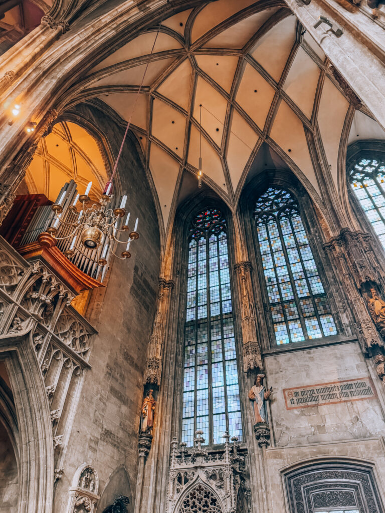 Detailed view of the vaulted ceiling and tall stained glass windows inside St. Stephen’s Cathedral in Vienna. The Gothic architecture features ribbed arches, ornate carvings, statues of saints, and a glimpse of the cathedral’s grand pipe organ.