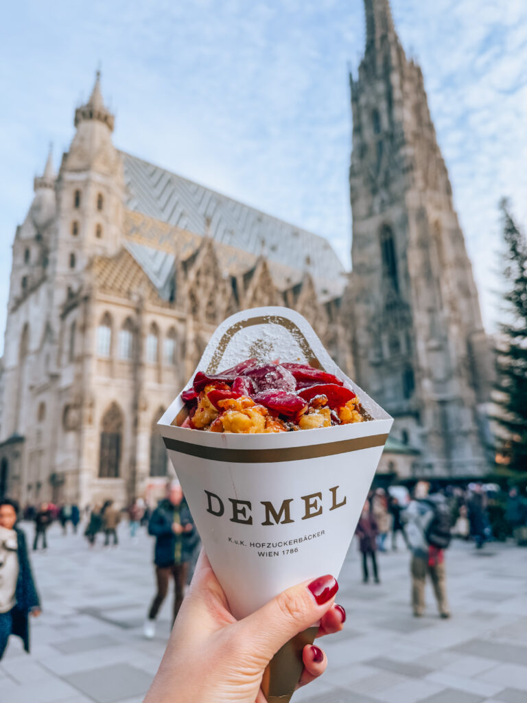 A hand holding a paper cone of Kaiserschmarrn topped with plum compote from Café Demel, with the iconic St. Stephen’s Cathedral (Stephansdom) in Vienna blurred in the background. The golden, caramelized shredded pancake contrasts with the historic Gothic church and bustling square behind.