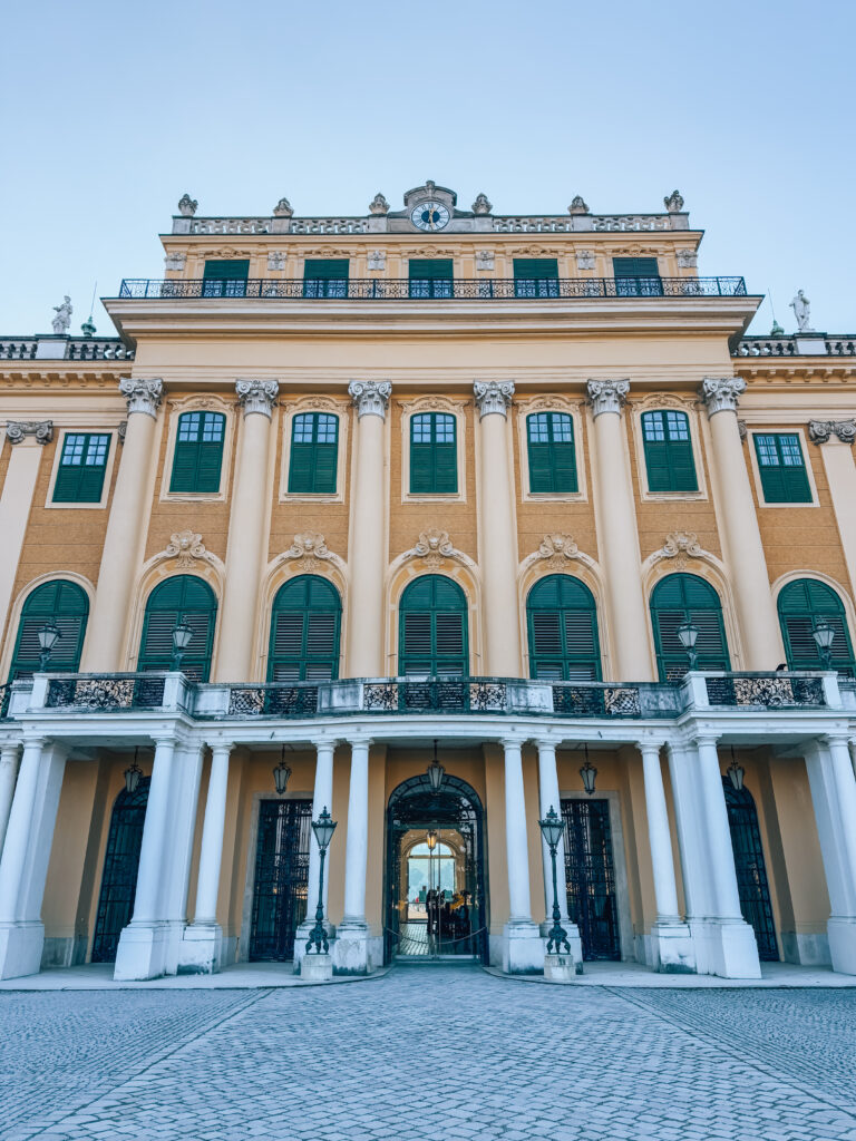 Front facade of Schönbrunn Palace in Vienna, showcasing its grand Baroque architecture with symmetrical green-shuttered windows, white columns, ornate statues, and an arched entrance framed by wrought iron gates