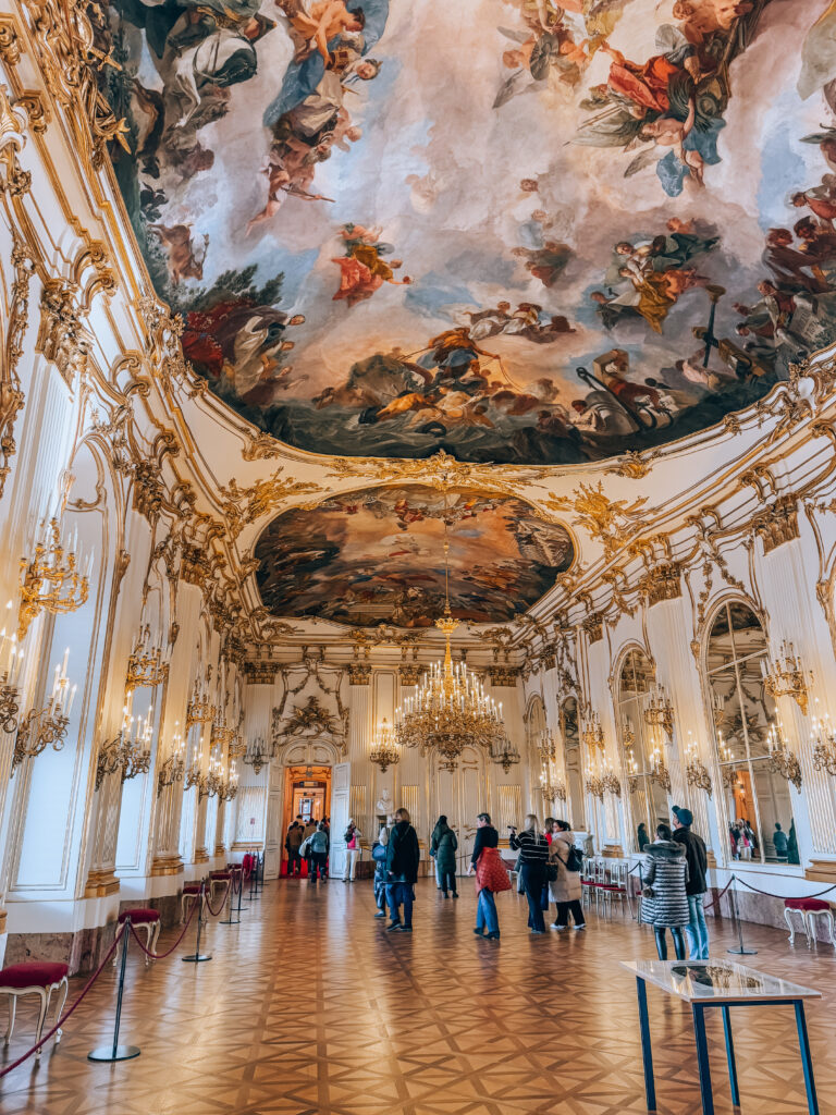 Interior of the State Apartments at Schönbrunn Palace in Vienna, featuring a stunning Baroque ceiling fresco, glittering chandeliers, mirrored walls, and golden stucco detailing. Visitors explore the grand ballroom's opulent space and parquet flooring.