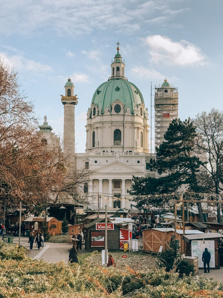 Stunning daytime view of Karlskirche (St. Charles's Church) in Vienna with its distinctive green dome and twin columns, surrounded by Christmas market stalls and visitors in the foreground, framed by winter trees and festive lights.