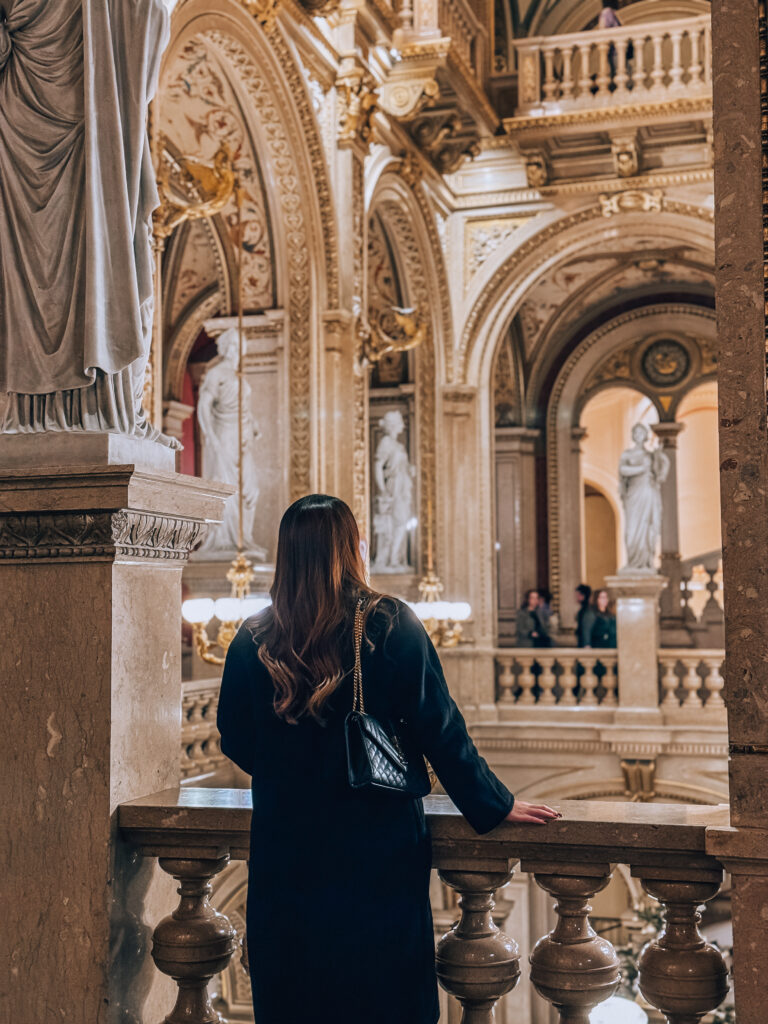 A woman in a dark coat stands on a marble balcony inside the Vienna State Opera, admiring the lavish interior filled with ornate arches, golden chandeliers, classical statues, and intricate architectural details.