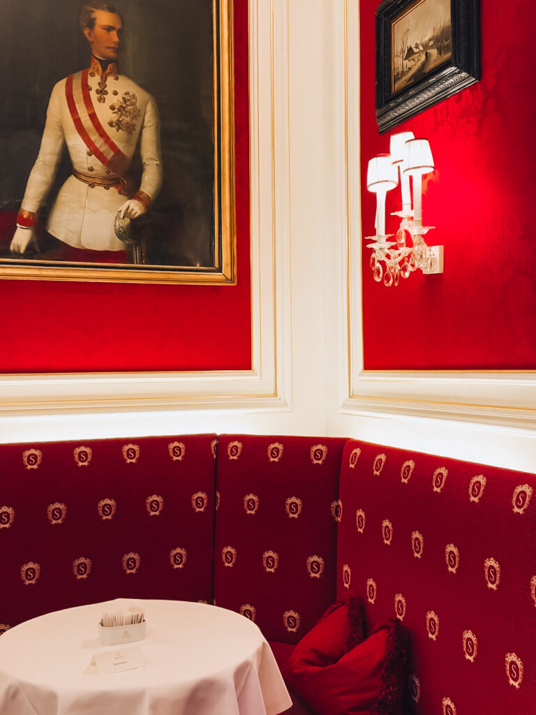 Elegant red velvet seating area at Café Sacher in Vienna, adorned with the signature gold “S” crest. A white tablecloth-covered table sits beneath a portrait of Emperor Franz Joseph, framed by cream-colored paneled walls and softly glowing crystal sconces.