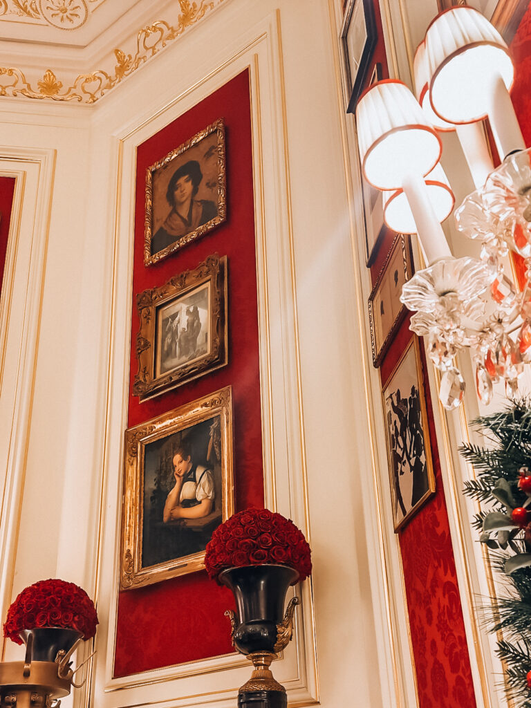 A richly decorated wall inside Café Sacher in Vienna, featuring ornate gold-framed portraits mounted on red velvet panels, flanked by crystal wall sconces and classical moldings. Two tall vases filled with red roses add to the luxurious and romantic ambiance.
