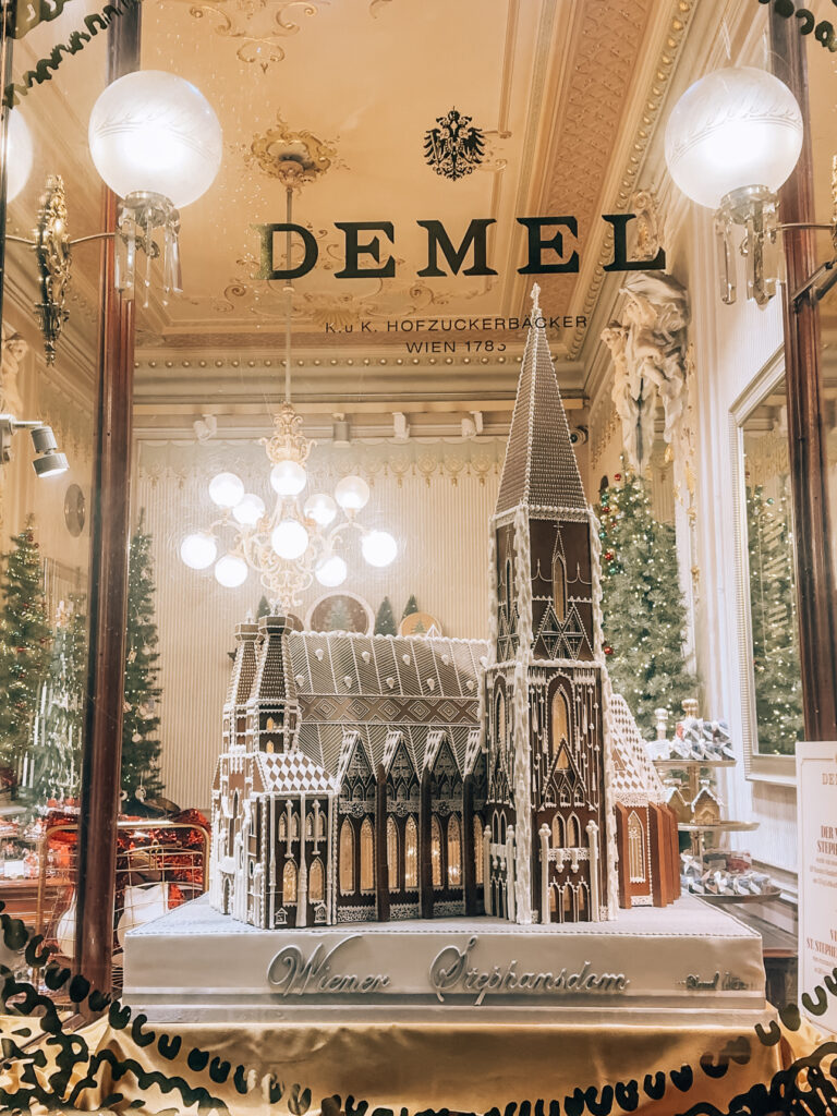 Holiday window display at Café Demel in Vienna featuring an intricate gingerbread replica of St. Stephen’s Cathedral (Wiener Stephansdom), surrounded by festive decorations and chandeliers inside the elegant historic patisserie.