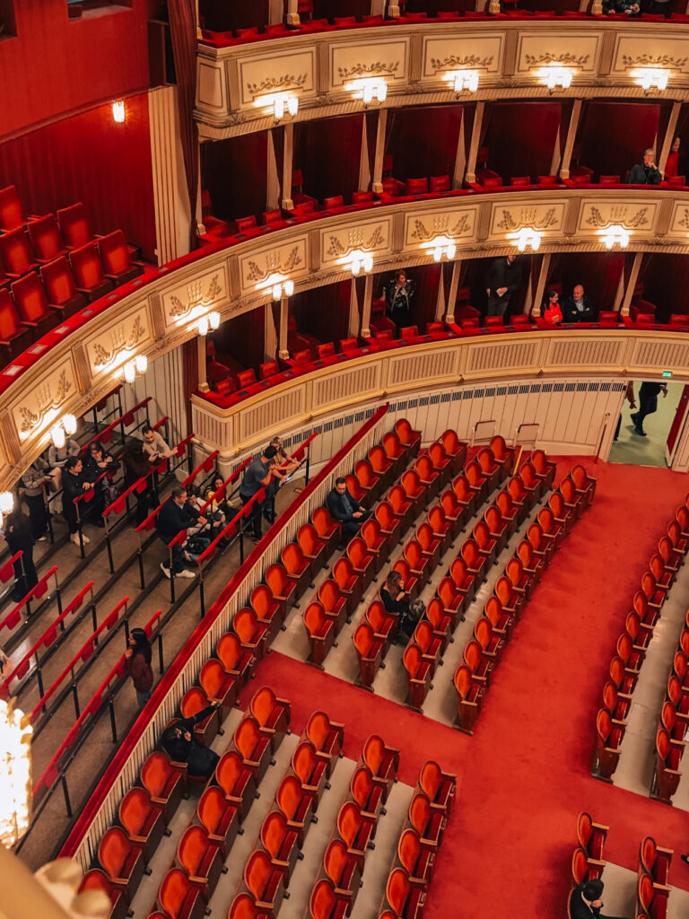 Interior view of the main auditorium at the Vienna State Opera, showing rows of plush red velvet seats, multiple tiers of ornate balconies with private boxes, and theatergoers beginning to take their seats before a performance.