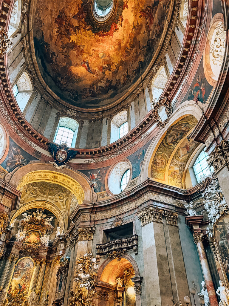 Interior view of St. Peter’s Church in Vienna, Austria, showcasing its ornate Baroque architecture, richly decorated altar, and the dramatic frescoed dome ceiling with vivid depictions of saints and angels illuminated by natural light from arched windows.