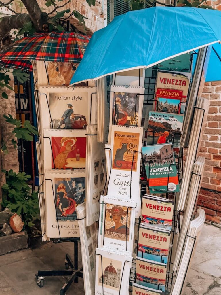 A display of retro style postcards and Venice guidebooks at Libreria Acqua Alta with colorful umbrellas shading the stand.