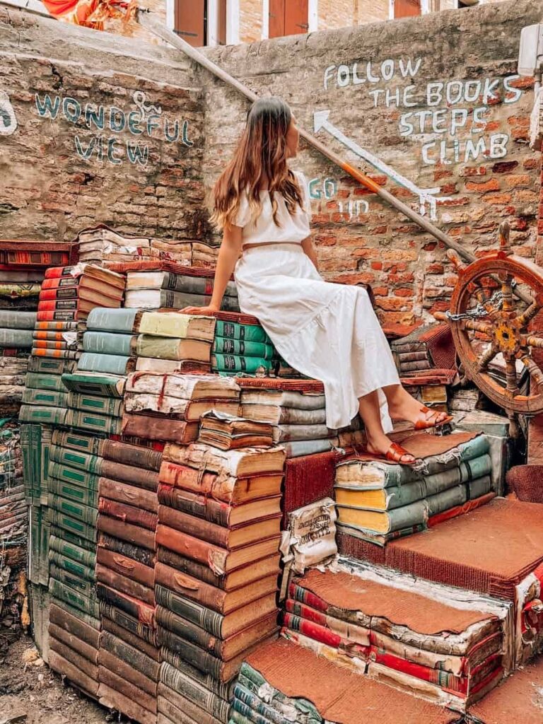 A woman sitting on the famous outdoor staircase made of old books at Libreria Acqua Alta with painted signs on the brick wall behind her.