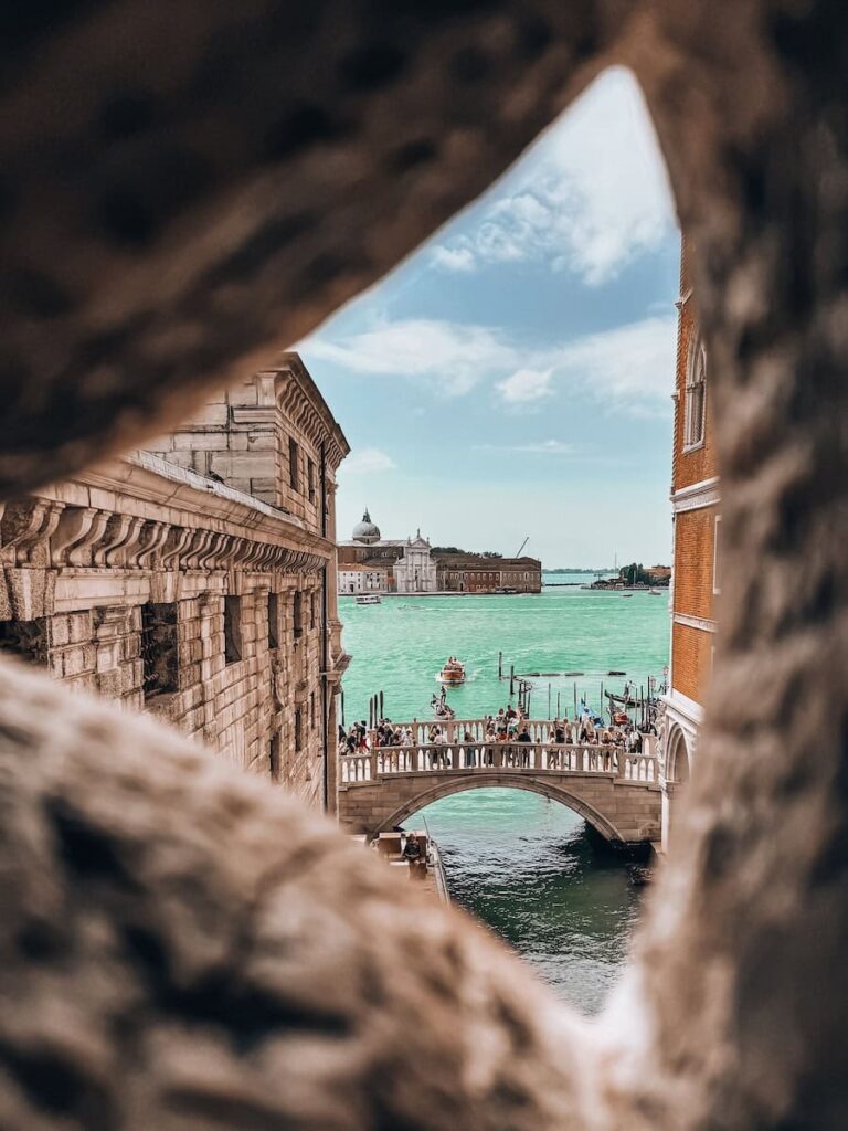 View of the Bridge of Sighs in Venice framed through a stone window, showing turquoise water, boats, and crowds on the bridge.