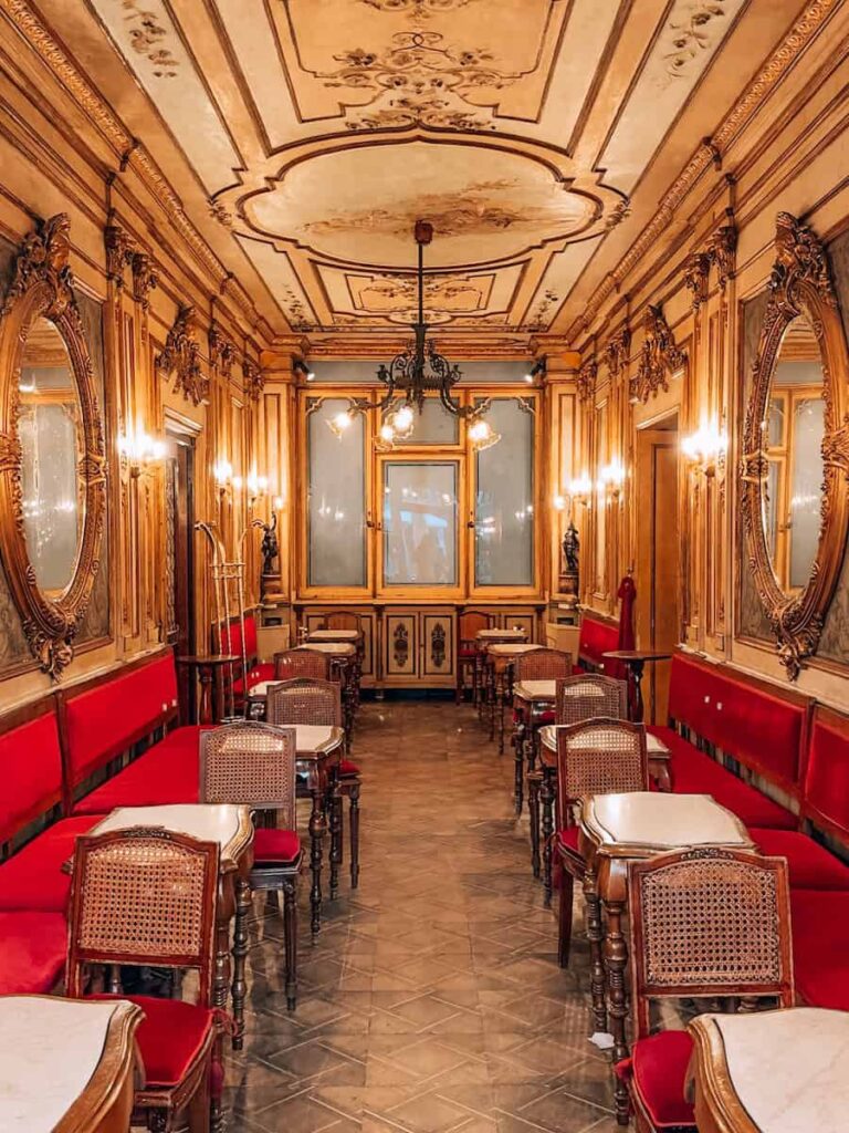 A long ornate dining room at Caffe Florian with red velvet seating, marble tables and detailed ceiling artwork.