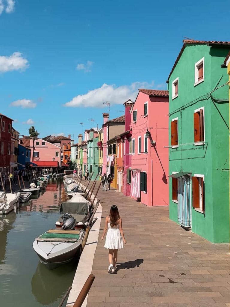 A woman in a white dress walking beside the canal in Burano surrounded by vibrant pastel houses and small boats.