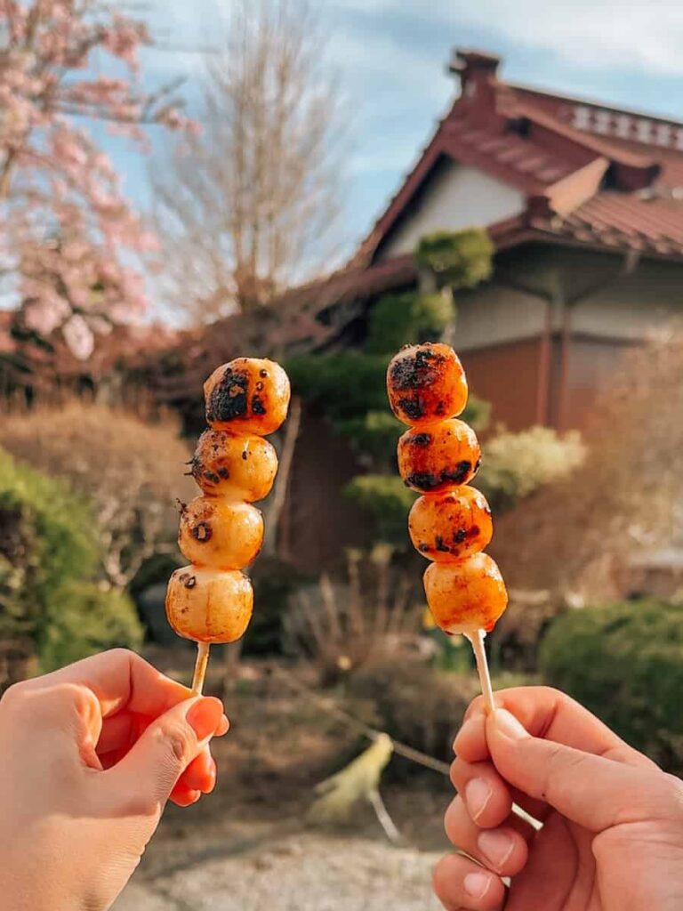 Two skewers of grilled dango held up outdoors with a traditional Japanese house and cherry blossoms in the background.