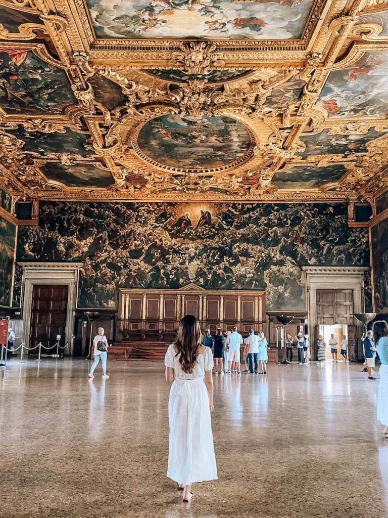 A woman in a white dress walking through the vast Great Council Chamber in the Doge’s Palace, surrounded by ornate gold ceilings and dramatic wall paintings.