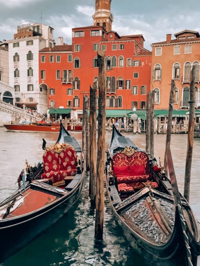 Two ornate gondolas with red velvet seats docked by wooden posts along the Grand Canal with colorful historic buildings in the background.