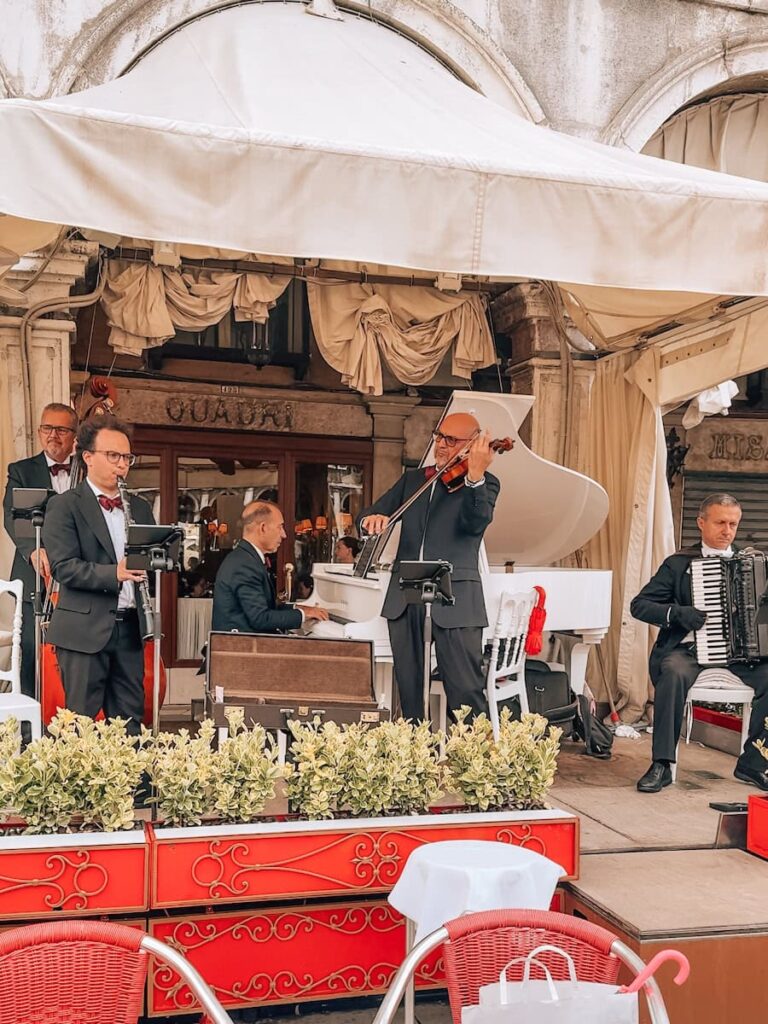 A live band performing at an outdoor cafe in Venice with musicians playing violin, piano, accordion and double bass in front of a historic building.