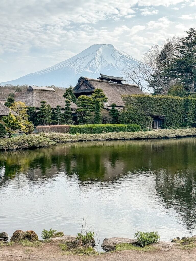 Scenic view of Mount Fuji reflected in a pond with traditional thatched-roof houses surrounded by greenery.