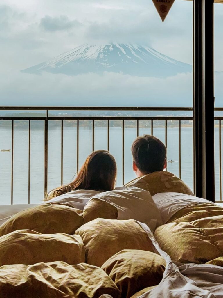 Couple relaxing in bed with panoramic views of Mount Fuji across Lake Kawaguchi from their ryokan room.