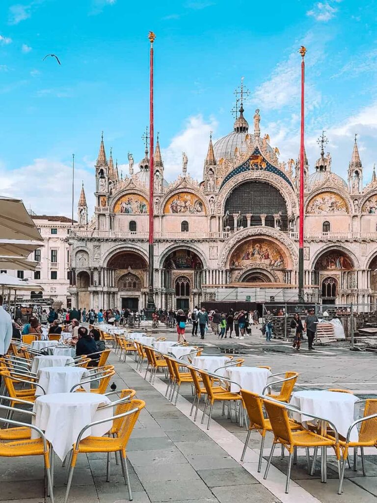 Rows of yellow cafe chairs and white tablecloths in Piazza San Marco with a clear view of the ornate facade and domes of St. Mark’s Basilica.