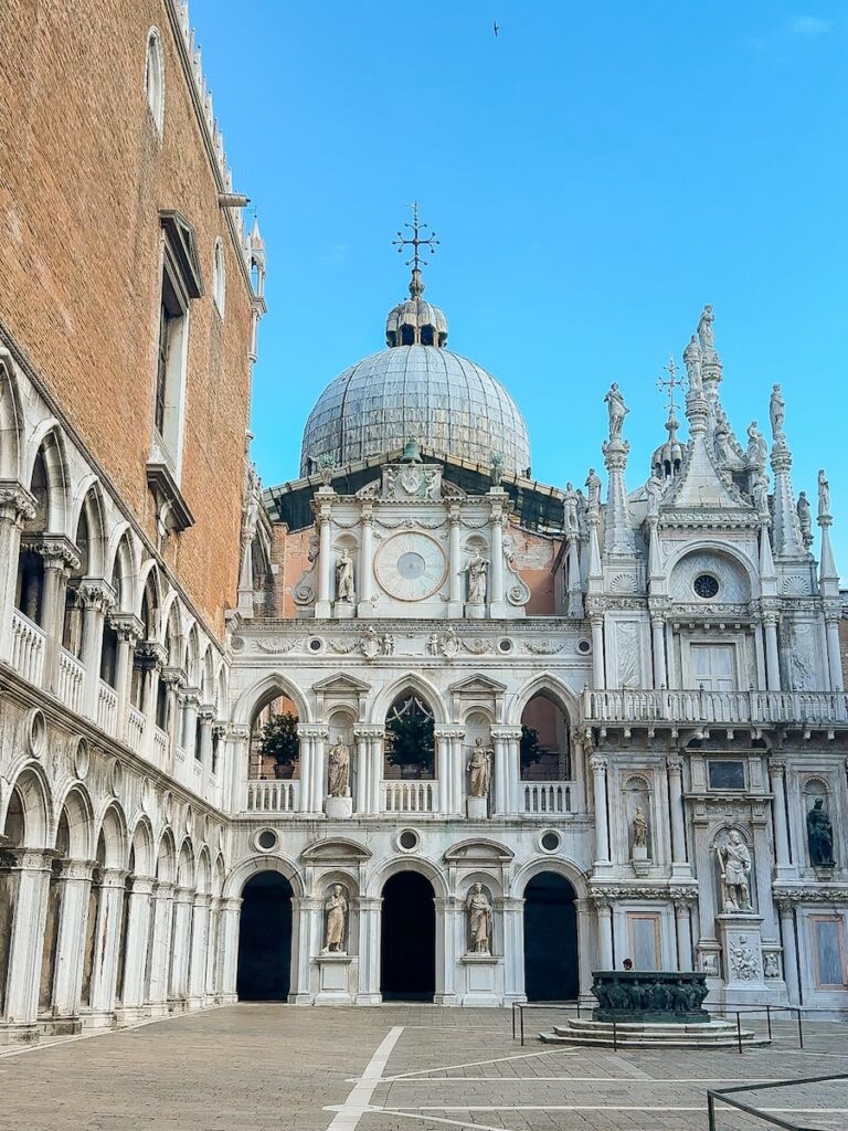 A quiet courtyard inside the Doge’s Palace featuring arched marble architecture, statues and the domed roof of St. Mark’s Basilica in the background.