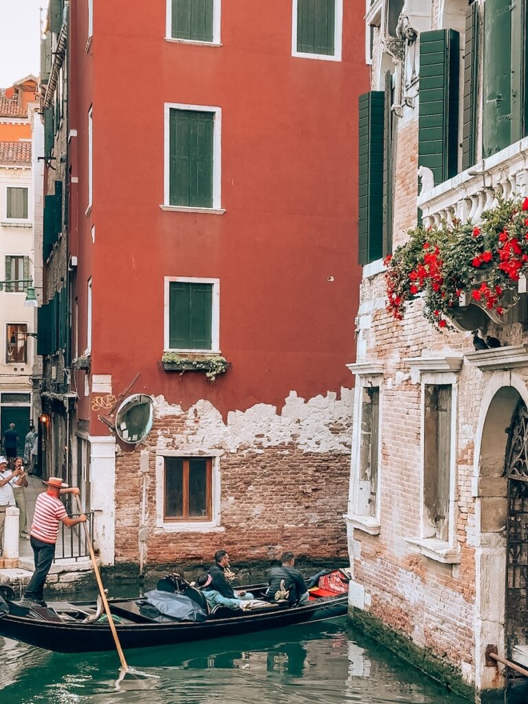 A gondolier steering a gondola past a red stucco building and a balcony with vibrant red flowers along a quiet canal in Venice.