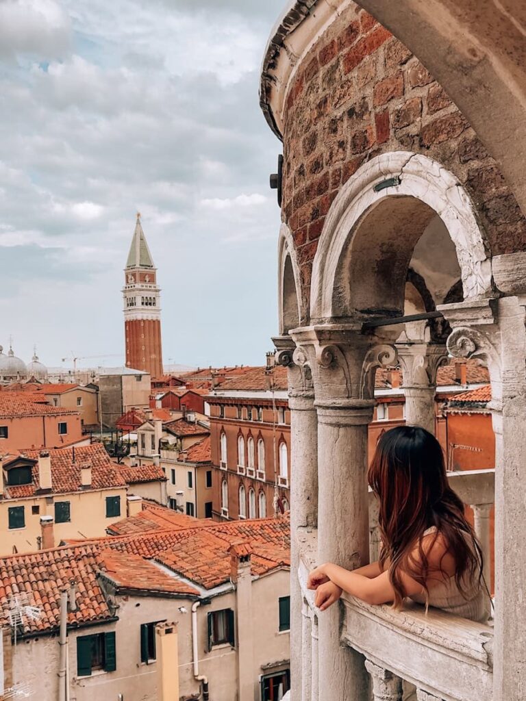A woman leaning on the stone balcony of the famous spiral staircase at Palazzo Contarini del Bovolo overlooking the terracotta rooftops of Venice.