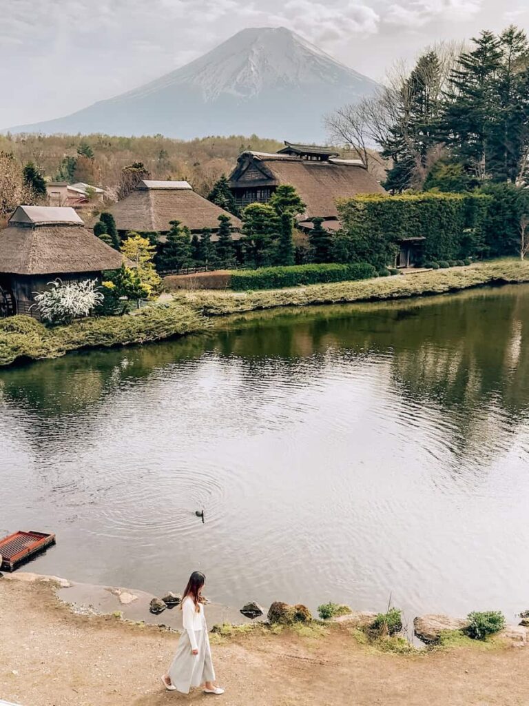 Woman walking along the pond at Oshino Hakkai with traditional thatched-roof houses and Mount Fuji in the background on a peaceful spring day.