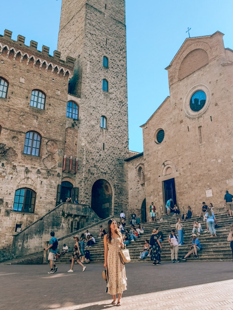 A woman in a floral dress stands in the main square of San Gimignano, Italy, holding a straw bag and sunhat, with the town’s medieval stone buildings, tower, and church steps in the background, bustling with visitors.