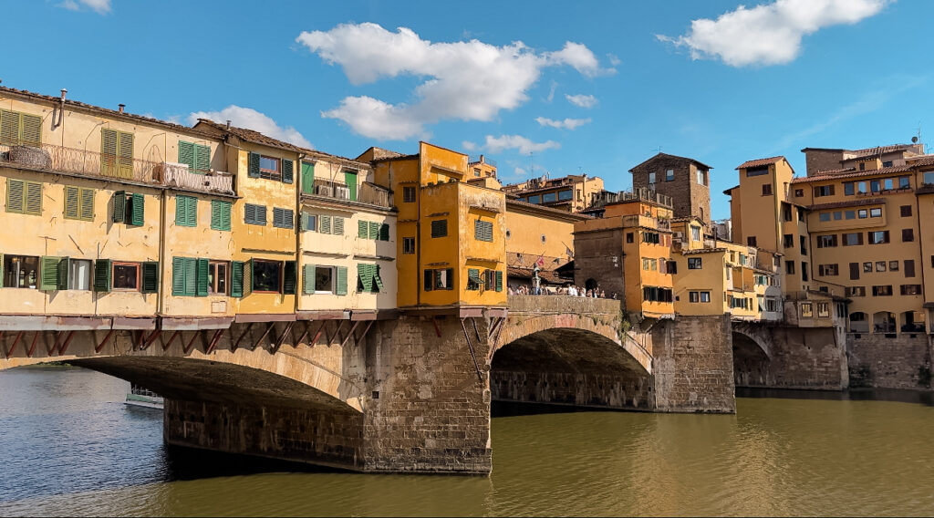 The Ponte Vecchio in Florence, Italy, a historic medieval stone bridge spanning the Arno River, lined with charming yellow and ochre buildings housing shops, with green shutters and a clear blue sky overhead.