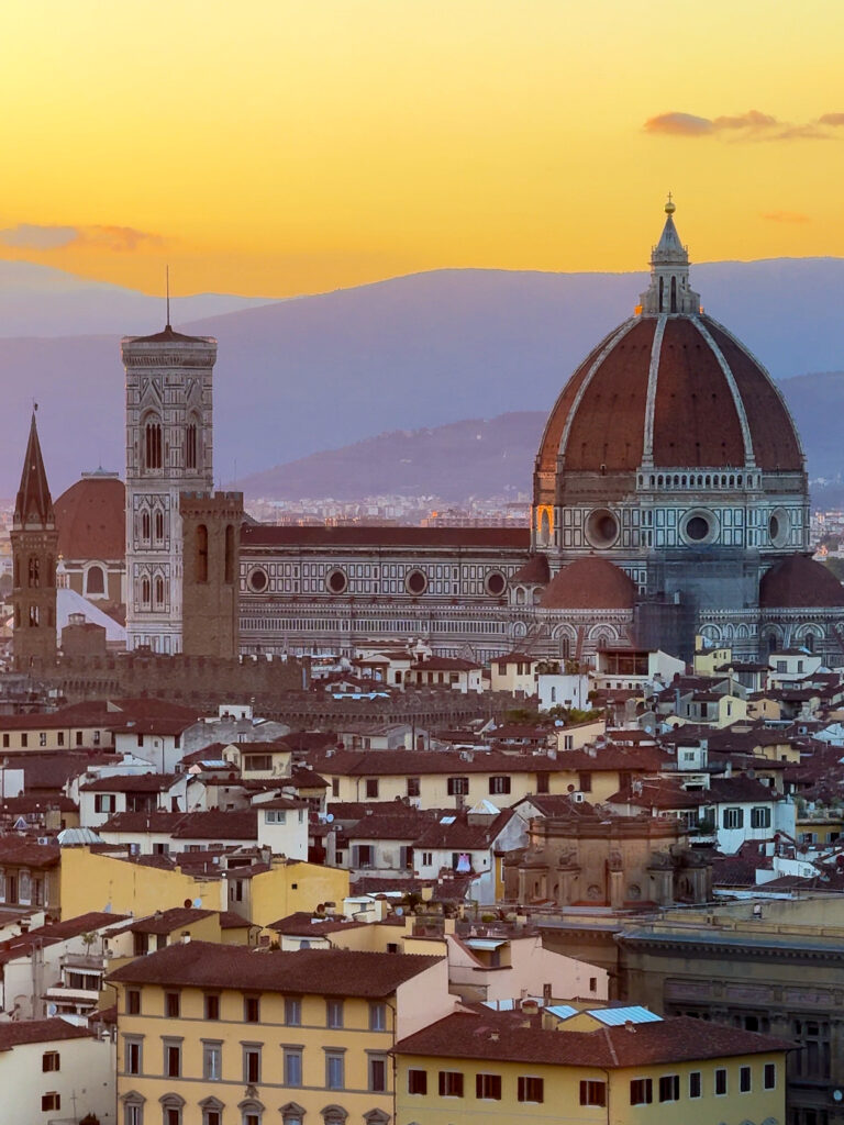 The Florence skyline at sunset, with the iconic red-tiled dome of the Cathedral of Santa Maria del Fiore and Giotto’s Bell Tower illuminated in golden light, set against rolling hills and a pastel-colored sky.