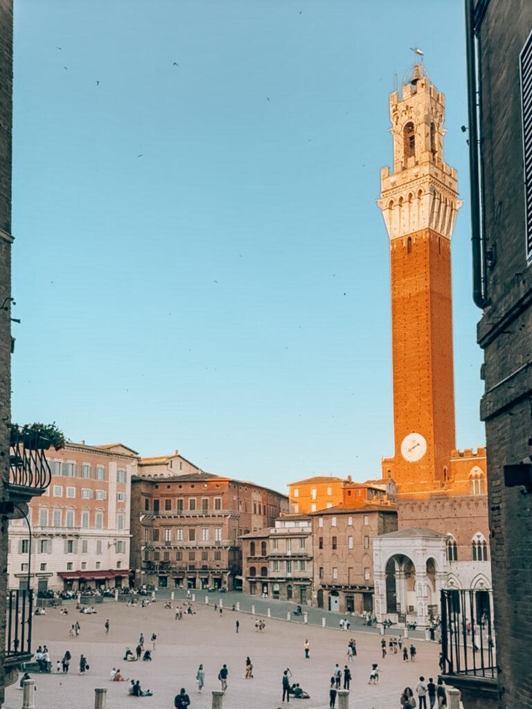 Piazza del Campo in Siena, Italy, with people scattered across the spacious square, surrounded by historic buildings, and the tall Torre del Mangia tower rising prominently against a clear blue sky.