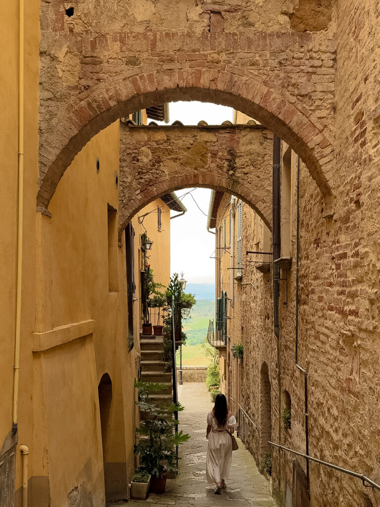 A woman in a flowing white dress walks down a narrow stone alley in Montepulciano, Italy, framed by rustic buildings and brick archways, with a glimpse of the countryside in the distance.