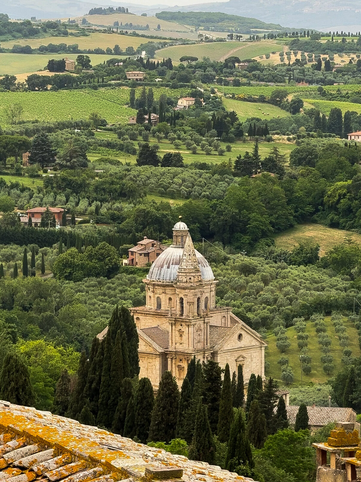 The Church of San Biagio in Montepulciano, Italy, with its striking Renaissance architecture and domed roof, nestled among cypress trees, vineyards, and rolling hills of the Tuscan countryside.