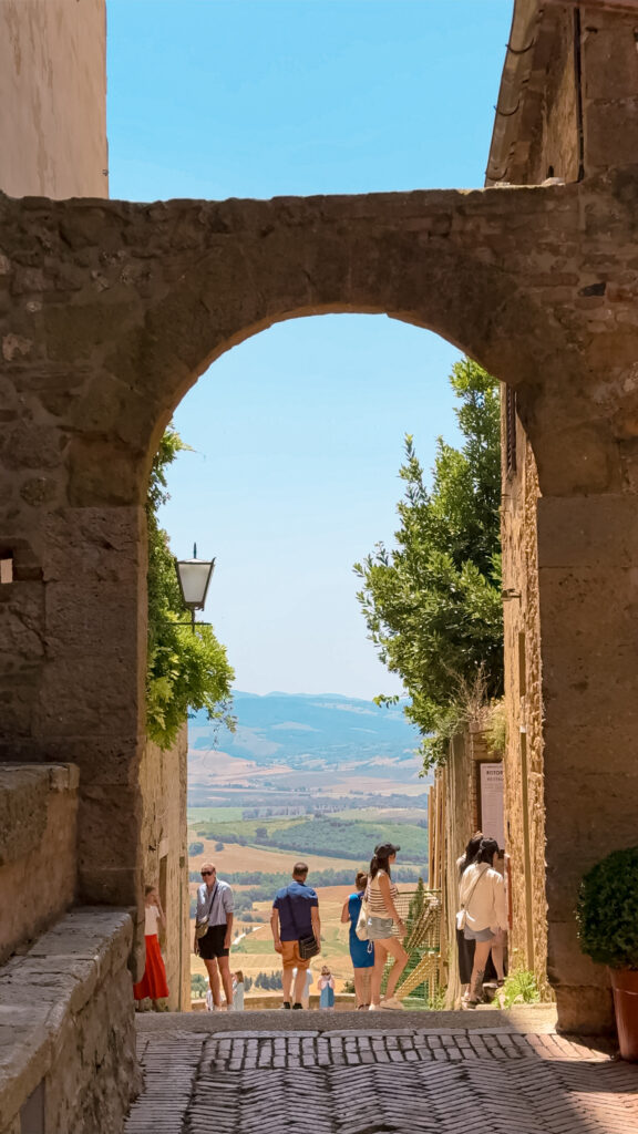 A stone archway framing a view of the Tuscan countryside, with visitors strolling along the cobblestone path and rolling hills visible in the distance under a bright blue sky.