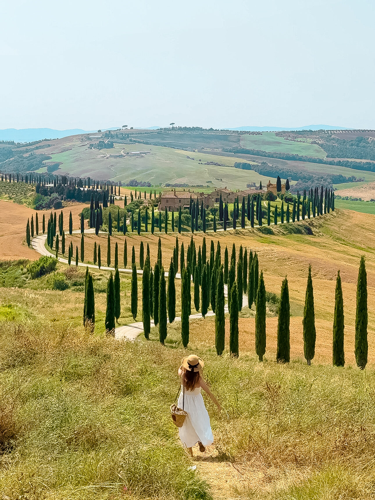 A woman in a white sundress and straw hat with a black ribbon walks through a grassy hill toward a winding cypress-lined road in the Tuscan countryside, with rolling hills and a rustic villa in the distance under a hazy summer sky.