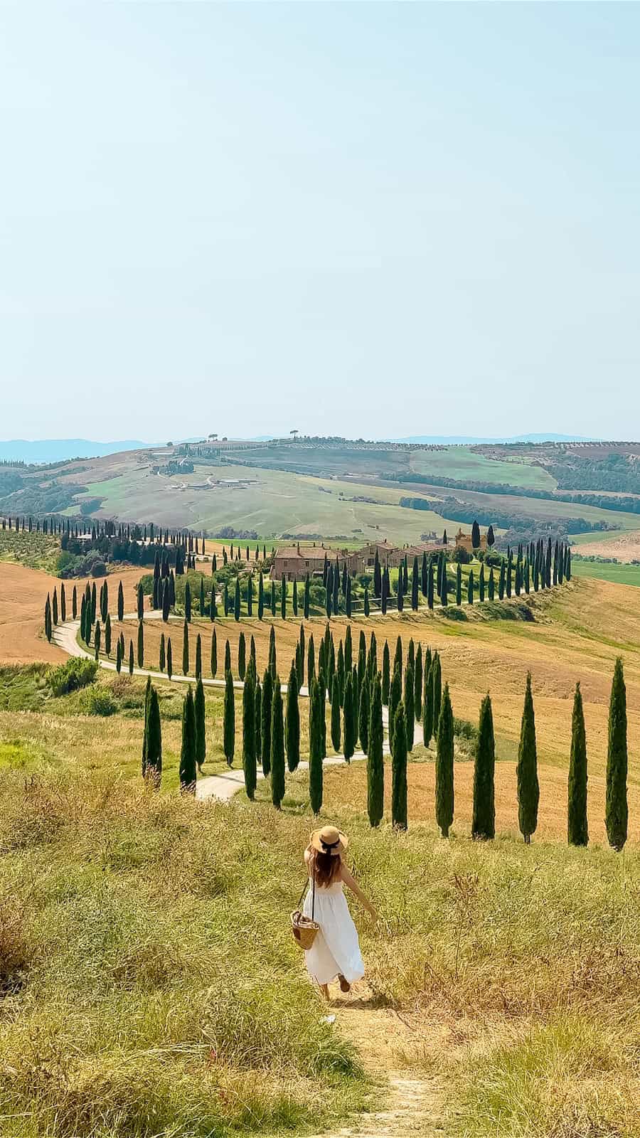 A woman in a white sundress and straw hat with a black ribbon walks through a grassy hill toward a winding cypress-lined road in the Tuscan countryside, with rolling hills and a rustic villa in the distance under a hazy summer sky.