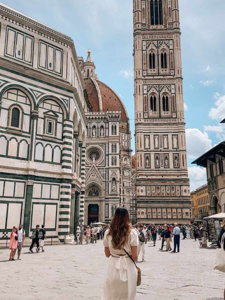 A woman in a white dress stands in the bustling Piazza del Duomo in Florence, Italy, gazing toward the Cathedral of Santa Maria del Fiore, with its intricate marble façade, the red-tiled dome, and Giotto’s Bell Tower rising against a partly cloudy sky.