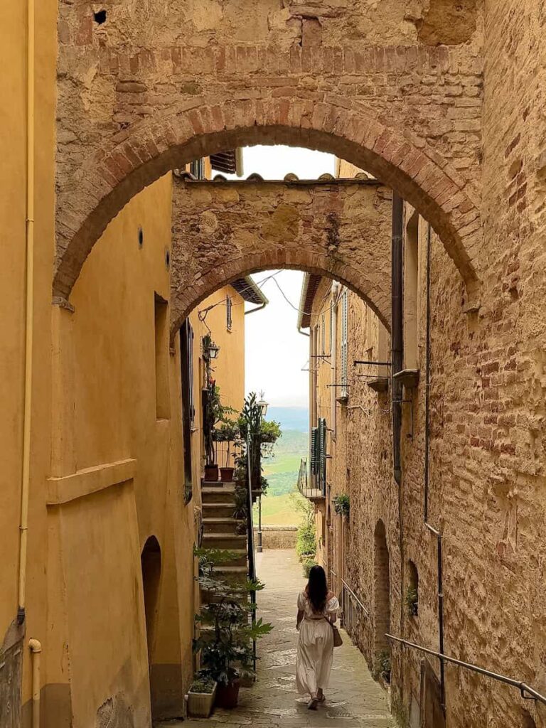 A woman in a flowing white dress walks down a narrow stone alley in Montepulciano, Italy, framed by rustic buildings and brick archways, with a glimpse of the countryside in the distance.