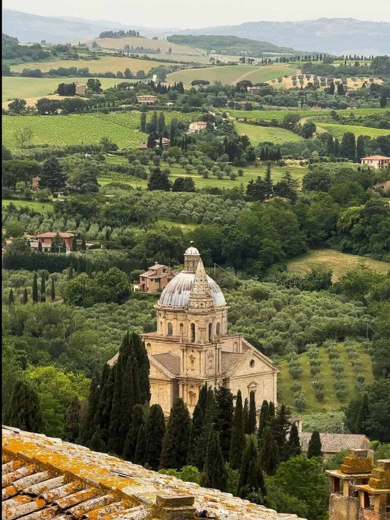 The Church of San Biagio in Montepulciano, Italy, with its striking Renaissance architecture and domed roof, nestled among cypress trees, vineyards, and rolling hills of the Tuscan countryside.