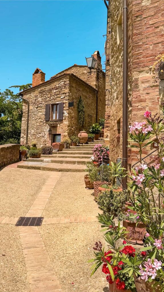A picturesque alley in a Tuscan village lined with potted flowers, leading to rustic stone houses with wooden shutters and terracotta roofs under a sunny sky.